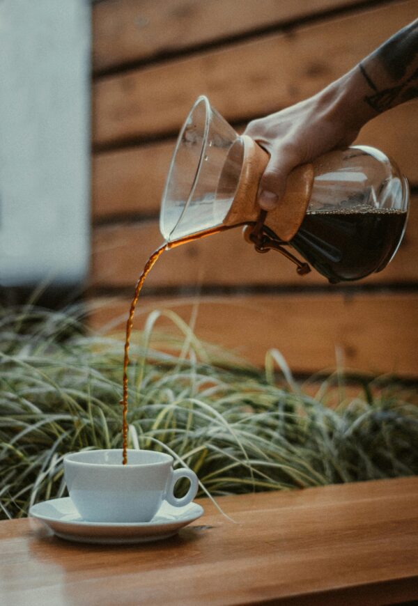 A hand pours freshly brewed coffee from a Chemex into a white cup in a cozy wooden setting.